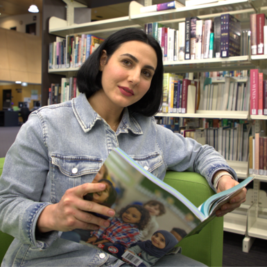 A woman with a dark bob hairstyle wearing a denim shirt is sitting on a green couch in a library reading a magazine.