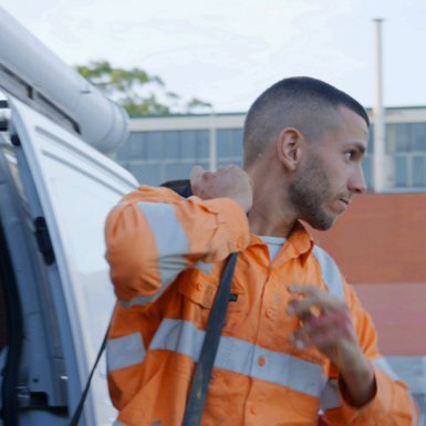 Plumbing student Joshua Presta in orange high vis shirt grabbing his tool bag from a white plumbing van in a carpark.