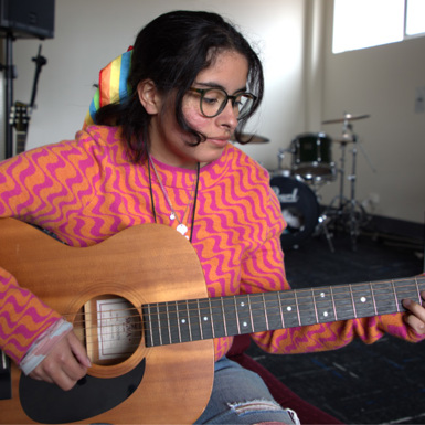 Songwriting and Music Production student Luca Munoz wearing a bright pink and orange patterned jumper and playing an acoustic guitar.