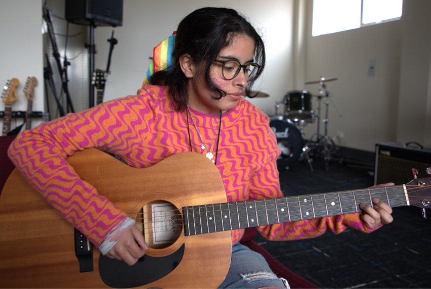 Songwriting and Music Production student Luca Munoz wearing a bright pink and orange patterned jumper and playing an acoustic guitar.