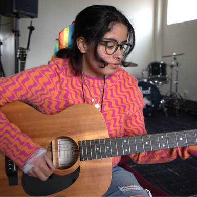 Songwriting and Music Production student Luca Munoz wearing a bright pink and orange patterned jumper and playing an acoustic guitar.