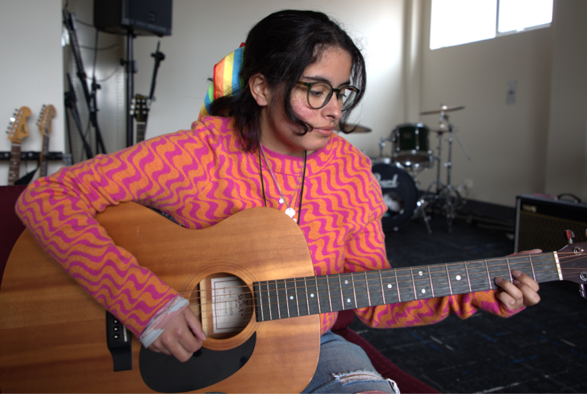Songwriting and Music Production student Luca Munoz wearing a bright pink and orange patterned jumper and playing an acoustic guitar.
