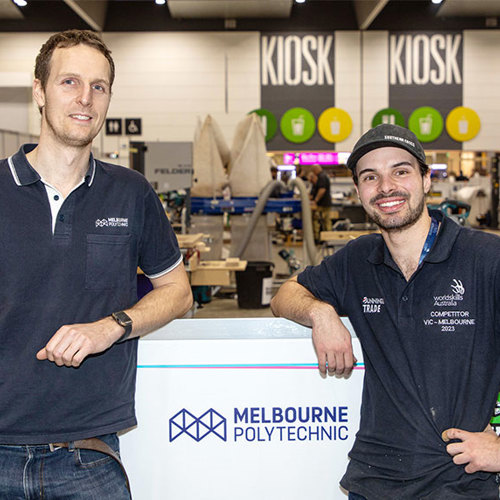 Two men standing together in front of the Melbourne Polytechnic booth at WorldSkills, looking towards the camera with friendly expressions.