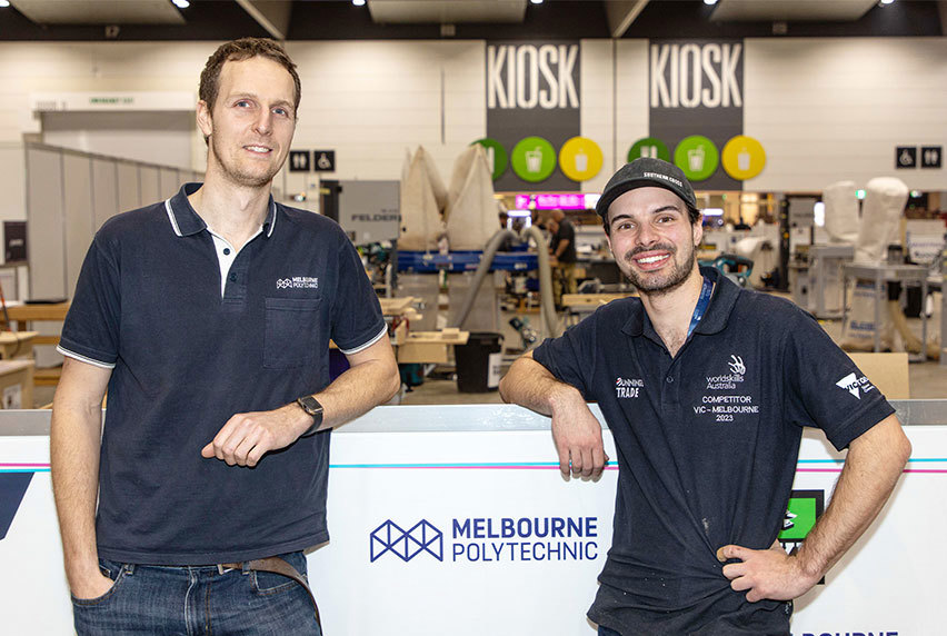 Two men standing together in front of the Melbourne Polytechnic booth at WorldSkills, looking towards the camera with friendly expressions.