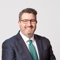 A well-dressed man in a suit and blue tie beams at the camera, showcasing a friendly and professional demeanor.