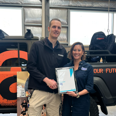 Carpentry teacher Lukas Page shakes hands with Margaret Mai from Selleys in front of a Carpentry Australia–branded ute, as they hold a framed scholarship certificate.