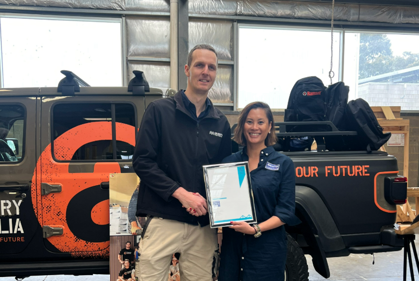 Carpentry teacher Lukas Page shakes hands with Margaret Mai from Selleys in front of a Carpentry Australia–branded ute, as they hold a framed scholarship certificate.