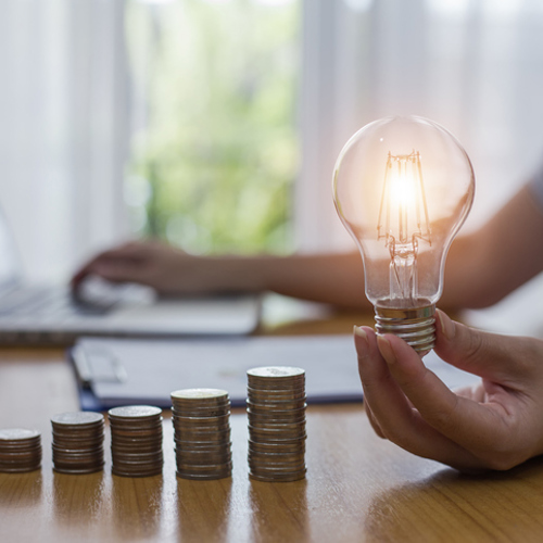 Stacks of coins arranged on a desk and a person holding a lit lightbulb while typing on a laptop