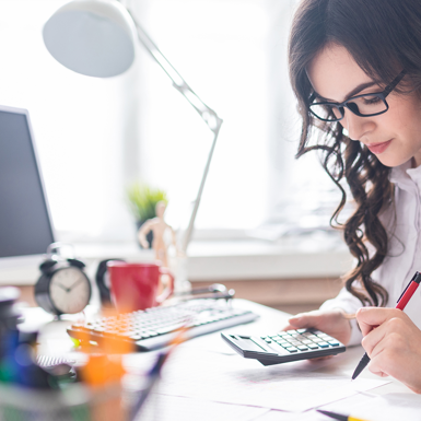 woman holding calculator, with paperwork in front of her and sitting at desk with computer