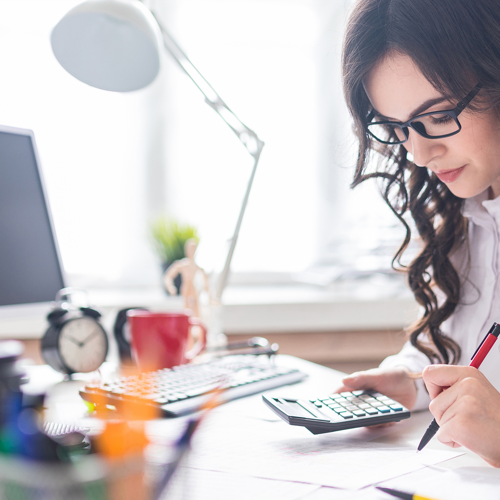 woman holding calculator, with paperwork in front of her and sitting at desk with computer