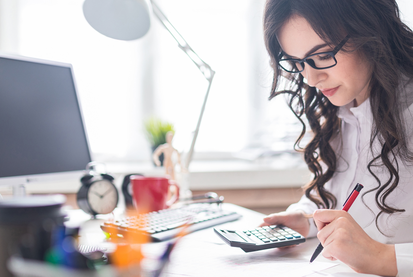 woman holding calculator, with paperwork in front of her and sitting at desk with computer
