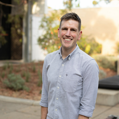A man wearing a light blue shirt, standing in a sunlit courtyard, smiling at the camera.