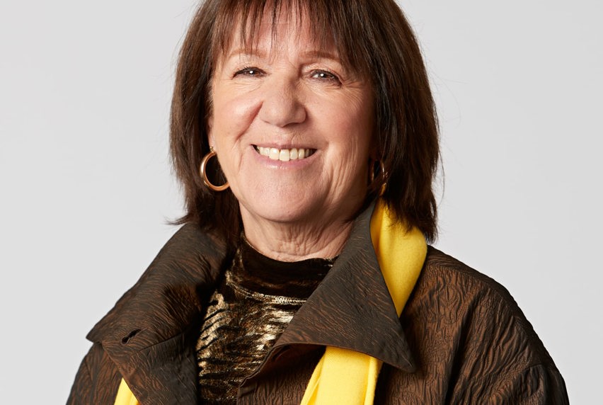 A woman wearing a brown jacket and a yellow scarf, standing confidently against a neutral background