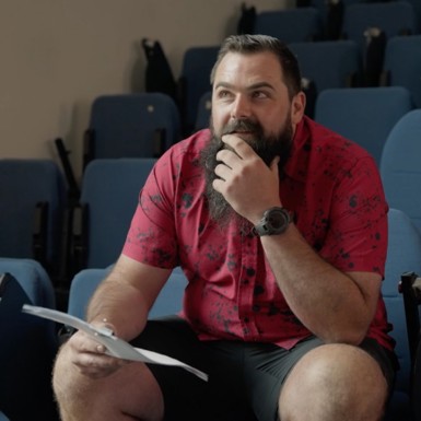 A bearded man wearing a red shirt, sits in a theater holding sheets of paper in front of him, in deep thought. 