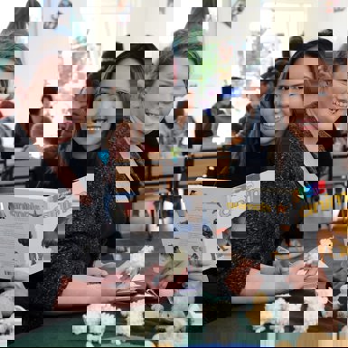  Two young women seated at a table smiling, surrounded by books and toys, engaged in a lively discussion.