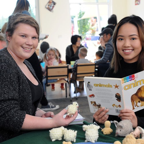  Two young women seated at a table smiling, surrounded by books and toys, engaged in a lively discussion.