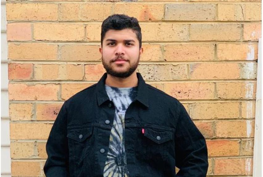 Portrait of Themiya in tie-dye t-shirt and black shirt standing in front of a brick wall