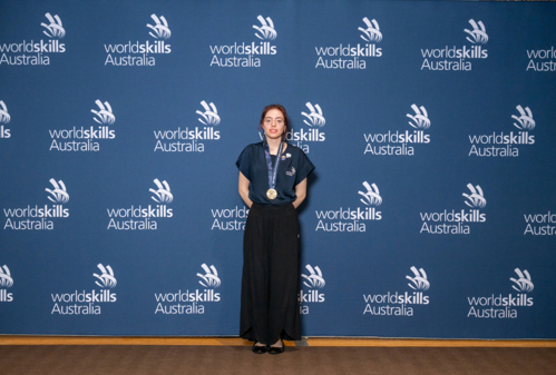 Taylah De Senna-Clark wearing WorldSkills uniform standing in front of a blue media wall with gold medal