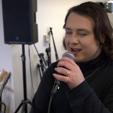 Dia Diamond (Zac Gelicrisio) rehearsing vocals in a Melbourne Polytechnic music studio, wearing a black turtleneck, manicured nails, gold rings and with dark shoulder-length hair. They are surrounded by studio equipment including speakers, guitars on a stand and a microphone stand.