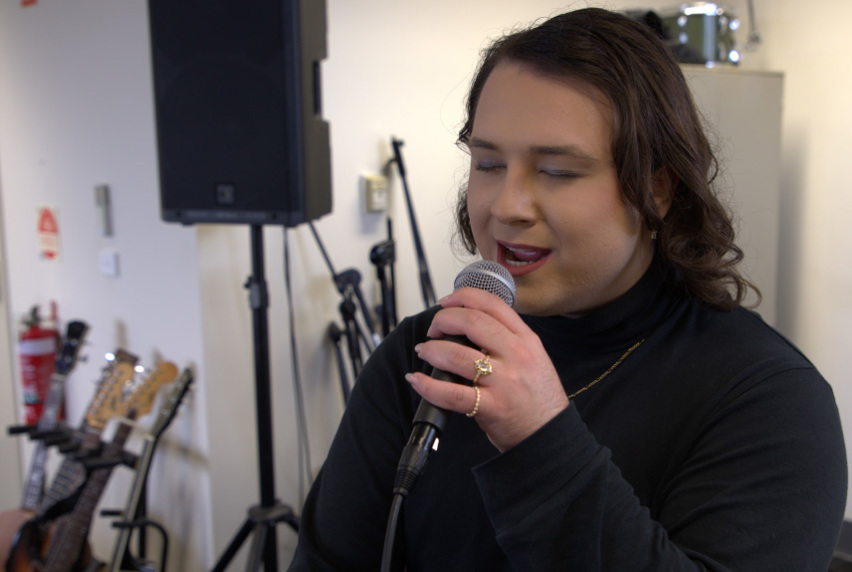 Dia Diamond (Zac Gelicrisio) rehearsing vocals in a Melbourne Polytechnic music studio, wearing a black turtleneck, manicured nails, gold rings and with dark shoulder-length hair. They are surrounded by studio equipment including speakers, guitars on a stand and a microphone stand.