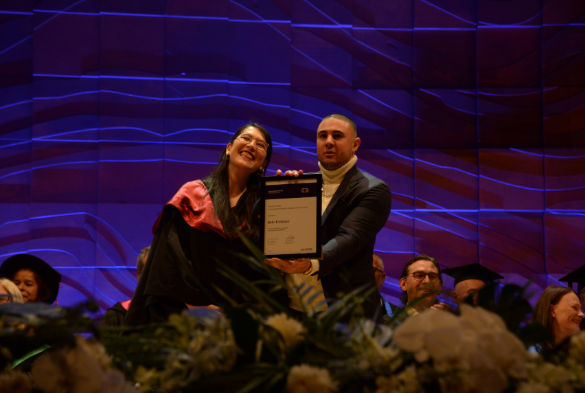 Two smiling people holding graduation certificate together on stage