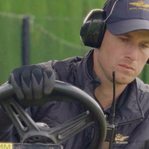 A man driving a ride-on lawn mower wearing hearing protection, gloves and a cap.