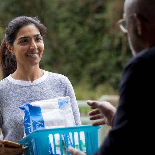 A community care volunteer delivering prescription medicine to a patient at their house. She is standing outside the door and passing it to the patient who is standing inside the house.