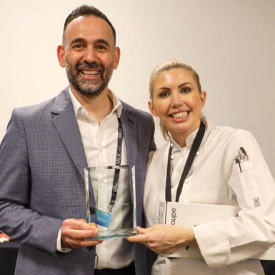 A man and a woman smiling, the woman is being presented with a glass trophy