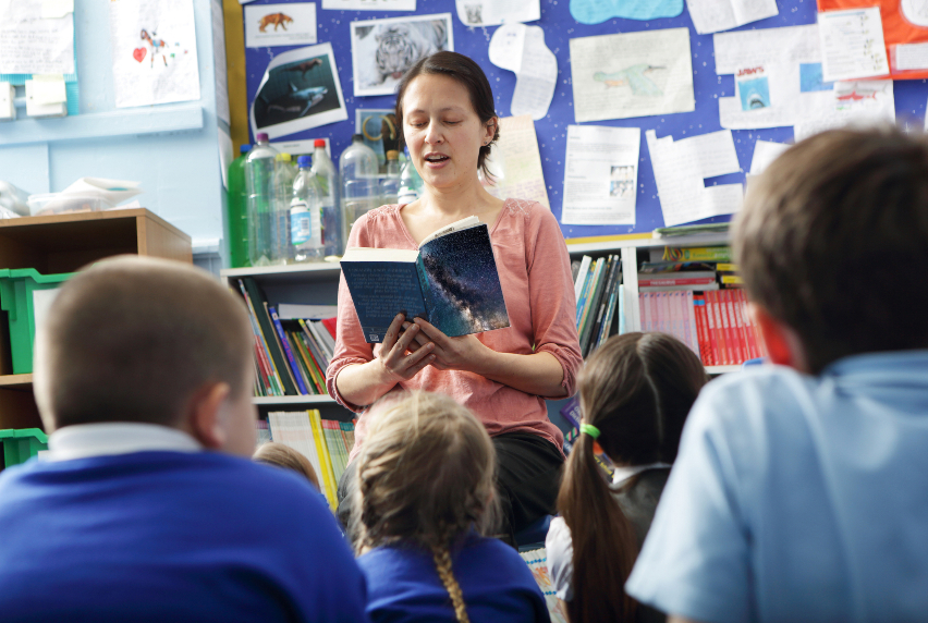 An educator reading to a group of primary school students, seated on the floor.