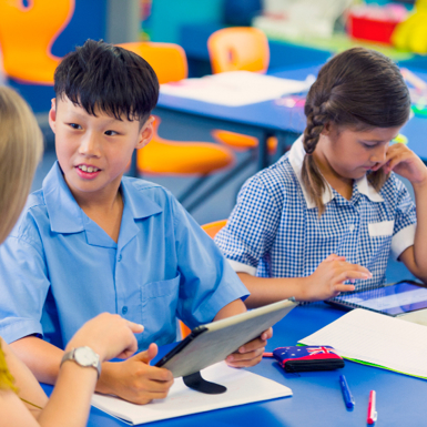 Two primary school students learning in a classroom with a smart tablet, assisted by a Teacher’s Aide.