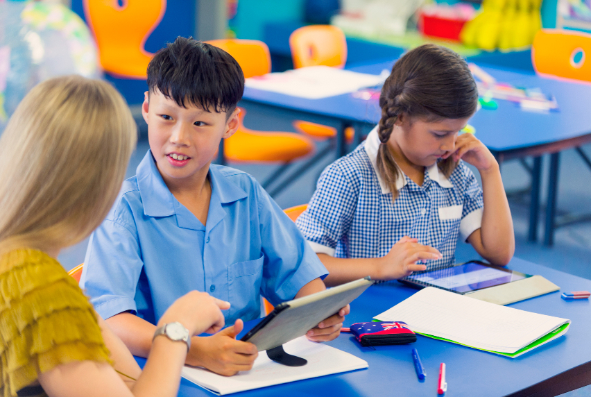 Two primary school students learning in a classroom with a smart tablet, assisted by a Teacher’s Aide.