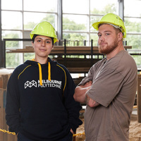 Woman and man posing confidently in a construction workshop wearing hardhats.