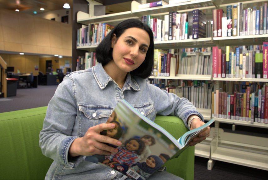 A woman with a dark bob hairstyle wearing a denim shirt is sitting on a green couch in a library reading a magazine.