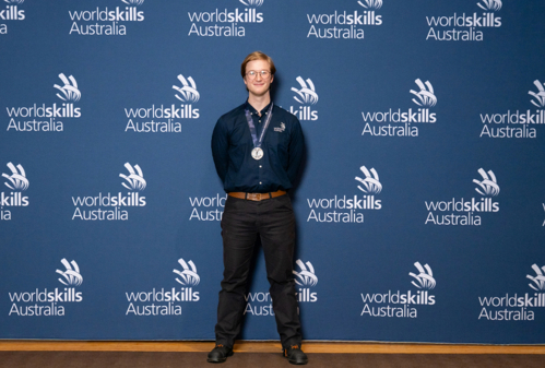 Paris Hermsen, Jewellery student wearing WorldSkills uniform in front of a blue media wall, smiling with silver medal.