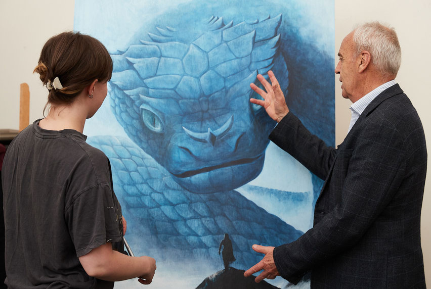 Teacher viewing student’s painting of a giant blue dragon towering over a small human on a mountain peak.