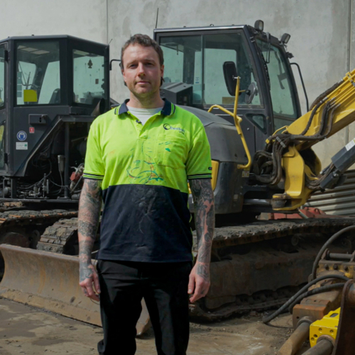  A young man wearing a high vis yellow t-shirt stands confidently in front of various construction equipment.