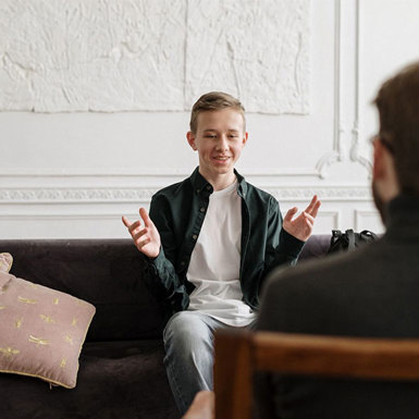 Young focused male seated on a couch, engaged in conversation with someone in front of him. 