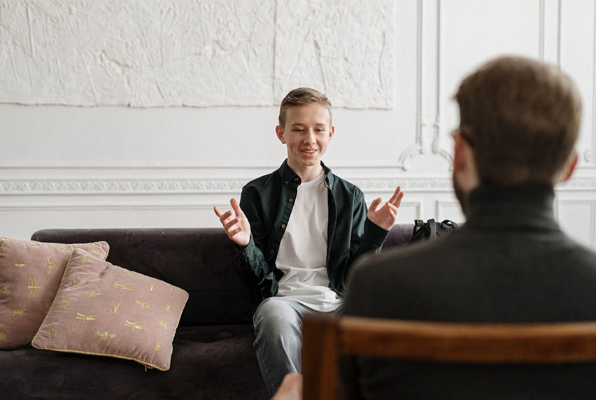 Young focused male seated on a couch, engaged in conversation with someone in front of him.