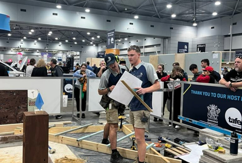 Andrew Millesi and William Hoffman looking at plan for Landscape Construction competition at WorldSkills. There is a barricade around the competition zone which contains wooden framing, tools and landscaping materials on the ground.