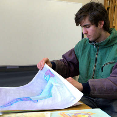 A student with dark brown short hair looking through a stack of colourful drawings at a table in a Visual Arts classroom.