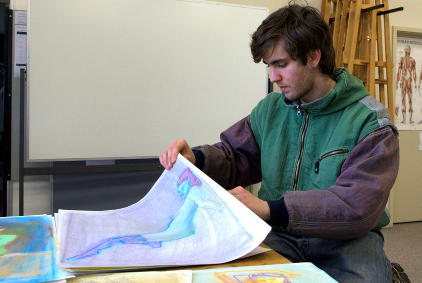 A student with dark brown short hair looking through a stack of colourful drawings at a table in a Visual Arts classroom.