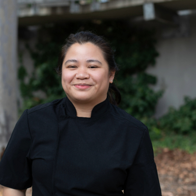 Woman wearing a black chef's uniform, smiling proudly with hair tied back, standing outside with trees and garden behind her.