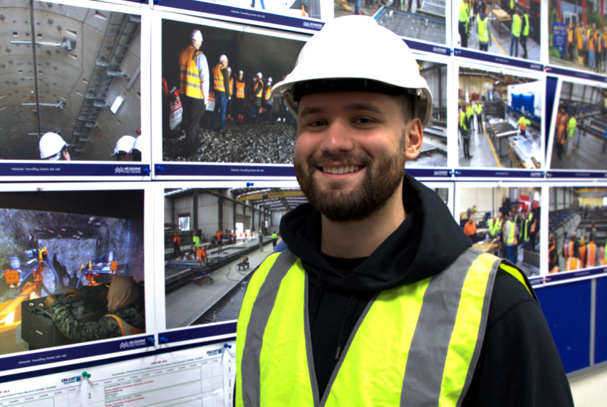 A male student wearing a black hoodie, yellow high vis vest and white hardhat standing in front of a display of construction site visit photographs.