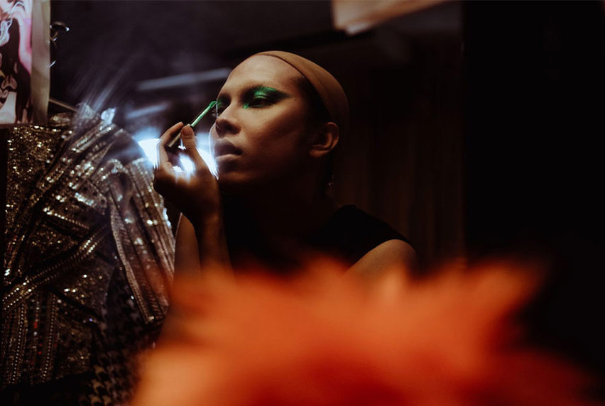 In mood lighting, a theatre actress applies green eyeshadow to her eyelids, while looking in a mirror.