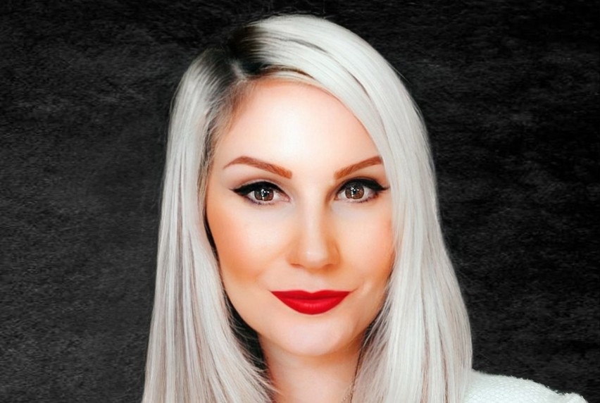 A woman with long white hair, striking red lipstick and wearing a white knitted top smiles at the camera wear