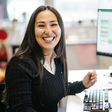 Image of a smiling brunette female, wearing a black V neck sweater. She is sitting in front of a computer.