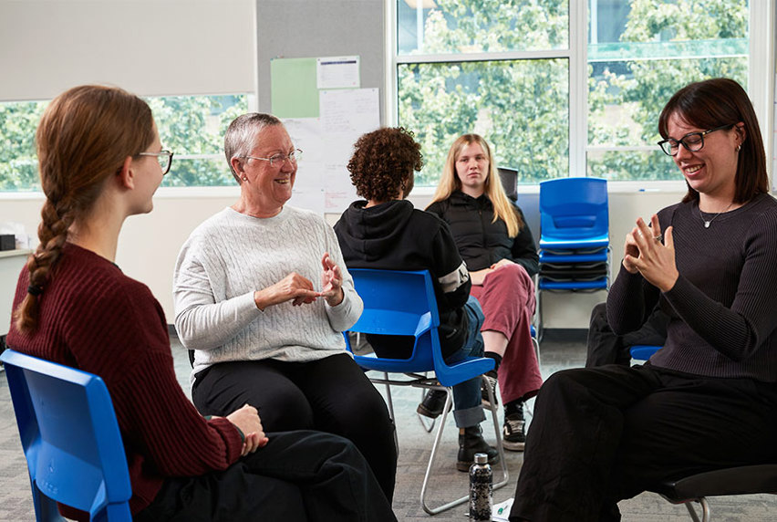 A diverse group of individuals sitting in chairs in a well-lit room, engaged in Auslan. 