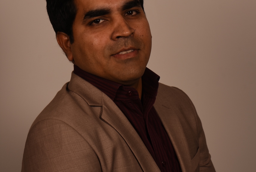 A man in a brown blazer and dark shirt poses at an angle, exuding sophistication.