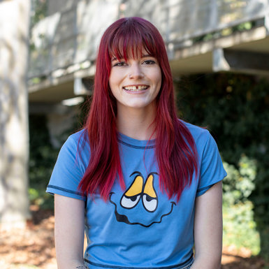 A woman with dyed vibrant red hair and a gap tooth smile, grinning at camera, wearing a blue tshirt with an illustrated face on it. She is standing in front of a garden and trees in the background.