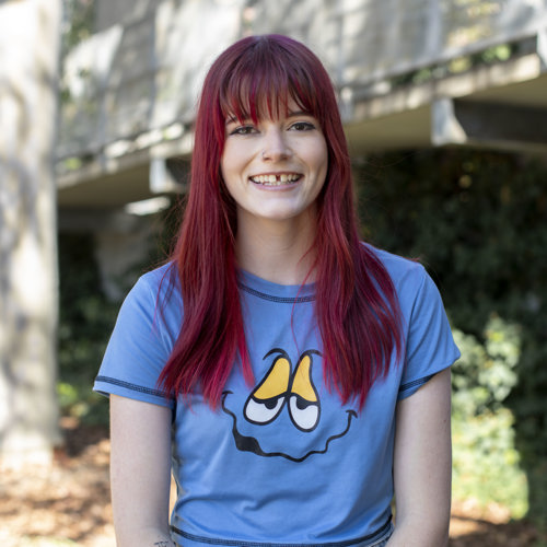 A woman with dyed vibrant red hair and a gap tooth smile, grinning at camera, wearing a blue tshirt with an illustrated face on it. She is standing in front of a garden and trees in the background.